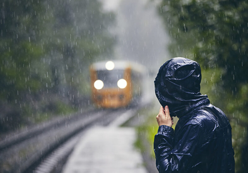 Man in drenched jacket standing on platform of railway station against train in heavy rain.