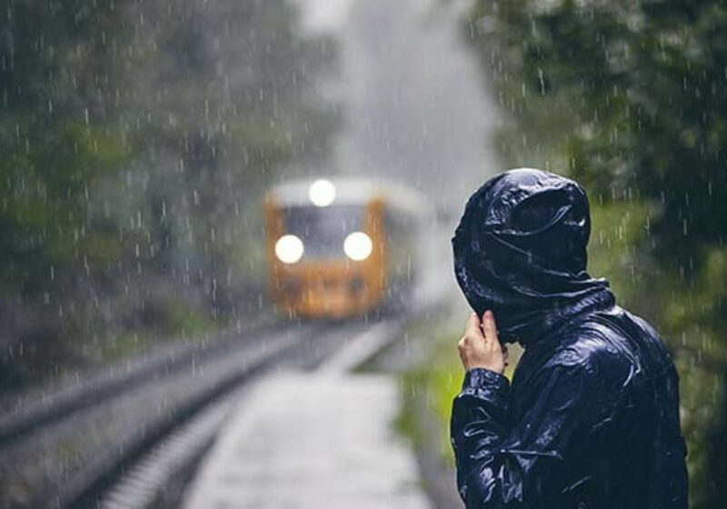 Man in drenched jacket standing on platform of railway station against train in heavy rain.